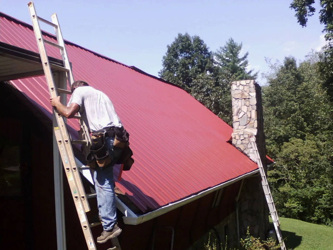 Roofer on a ladder beside a red metal roof and stone chimney — Soffit & Fascia Repair by Roofing Brothers in Meriden